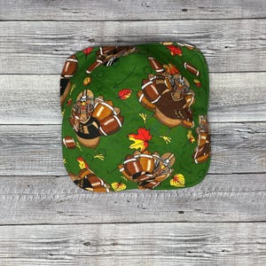 May include: Green fabric bowl with a Thanksgiving football theme. The bowl features turkeys wearing football helmets and holding footballs. The bowl is decorated with leaves and footballs.