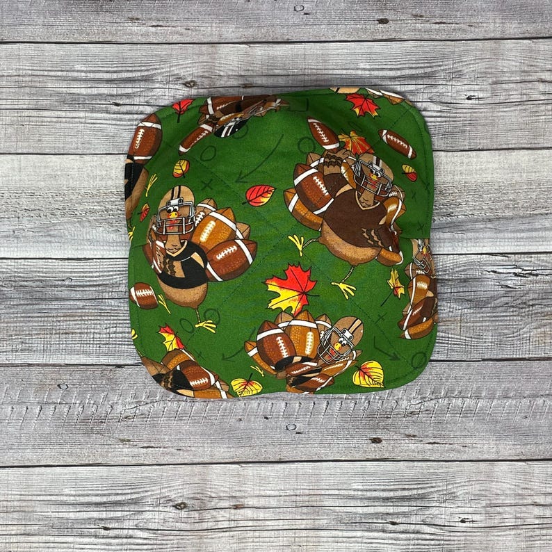 May include: Green fabric bowl with a Thanksgiving football theme. The bowl features turkeys wearing football helmets and holding footballs. The bowl is decorated with leaves and footballs.