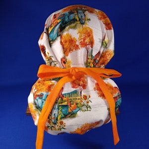 May include: A white fabric scrub hat with a fall-themed print of vintage trucks, trees, and flowers. The hat is tied with a bright orange ribbon.