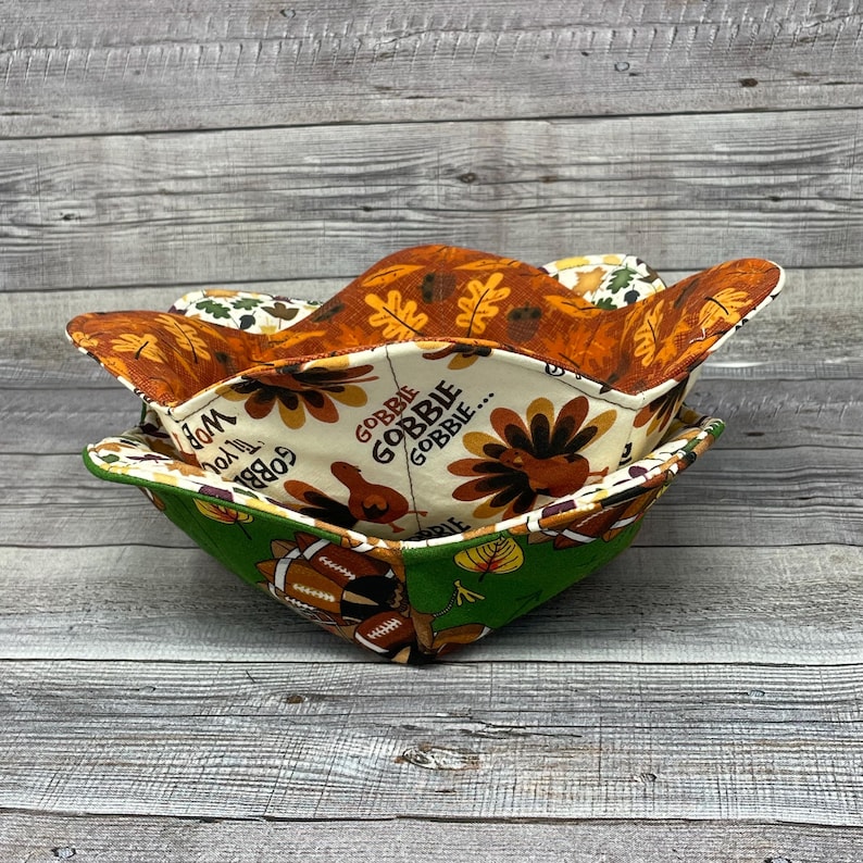 May include: Two fabric bowls with Thanksgiving themes. The top bowl is orange and brown with a leaf pattern. The bottom bowl is green with a football and turkey pattern. The text on the bowl reads "Gobble Gobble Gobble..."
