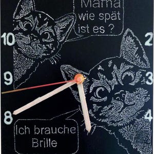 May include: Black square clock with white cat illustrations and hands. The clock face has the numbers 1-12. Text bubbles say "Mama wie spät ist es?" and "Ich brauche Brille". The clock hands are at 8:00. The clock is a decorative item.