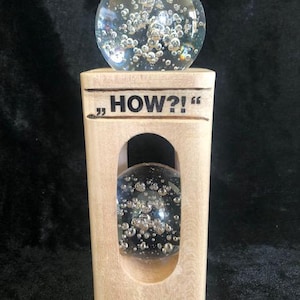 May include: A light-coloured wooden block sculpture featuring the text "HOW?!" engraved on the front. The block has a vertical oval cutout containing a clear glass sphere with internal bubbles, and a second identical glass sphere sits on top.