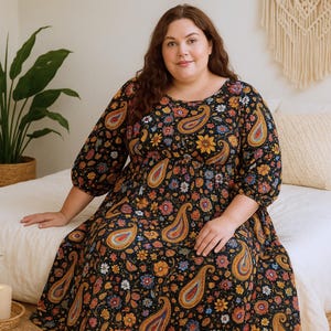 Peut inclure: Robe noire à manches longues ornée d'un motif cachemire coloré avec des touches d'orange, de jaune et de bleu. La robe a un col rond et une taille froncée. La personne est assise sur un lit.