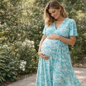 Puede incluir: Vestido de maternidad azul claro con un estampado de olas blancas. El vestido tiene mangas cortas y un diseño cruzado. La mujer está de pie al aire libre, rodeada de vegetación, y se sujeta suavemente el vientre. El vestido es largo.