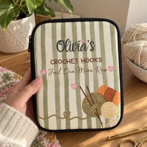 Puede incluir: Un estuche rectangular con cremallera y un patrón de rayas verticales en blanco y verde salvia. El texto dice Olivia's CROCHET HOOKS Just One More Row. Incluye ilustraciones de ovillos de lana, ganchillos y pequeños corazones rosas.