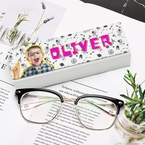 May include: Black and gold-rimmed eyeglasses with a personalized case. The case features the name "OLIVER" in pink letters, a photo of a child, and a pattern of astronauts and space-themed elements. The glasses are on a white surface.