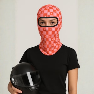 Può includere: Un passamontagna a scacchi rossi e rosa con un'apertura nera per gli occhi e piccoli disegni a stella bianchi. La persona indossa una maglietta nera e tiene in mano un casco da moto nero.