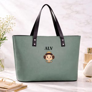 Puede incluir: Bolso tote verde salvia con asas negras y un gráfico de dibujos animados. La bolsa presenta las iniciales "ALV" encima del personaje de dibujos animados. La bolsa está sobre una superficie de mármol.