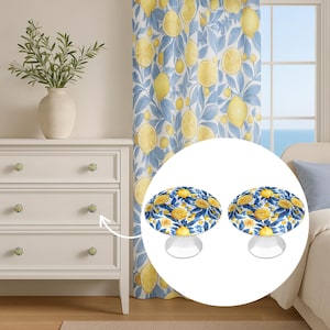 May include: Two mushroom-shaped cabinet knobs with a lemon and leaf design. The knobs feature a white base with yellow lemons and blue leaves. They are set on a white dresser with matching knobs. A curtain with a lemon pattern is visible in the background.