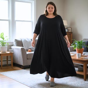 May include: A black, loose-fitting dress with a round neck and three-quarter length sleeves. The dress falls to the ankles. The person is wearing grey slip-on shoes. The setting is a bright living room with neutral tones and a window.