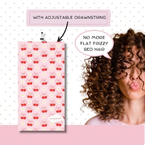 Puede incluir: Una máscara de dormir de satén rosa con un patrón de cerezas y flores. La máscara tiene un cordón ajustable y el texto "Le curly dreamin'" está impreso en la parte inferior.