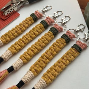 May include: Five handmade macrame keychains with a yellow, pink, white, and green color scheme. Each keychain has a silver metal key ring.