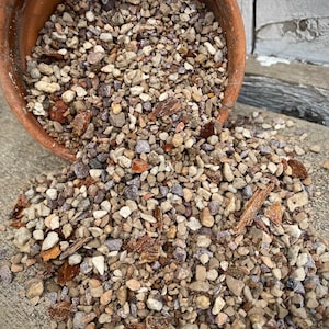 Könnte beinhalten: Ein Terrakotta-Topf gefüllt mit einer Mischung aus kleinen braunen, grauen und weißen Steinen. Der Topf ist umgekippt und die Steine fallen auf eine graue Oberfläche.