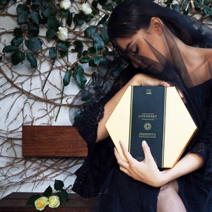 May include: A woman in a black dress and veil holds a hexagonal box with the text "LIFEHONEY" and a sun graphic. The box is gold with black accents.