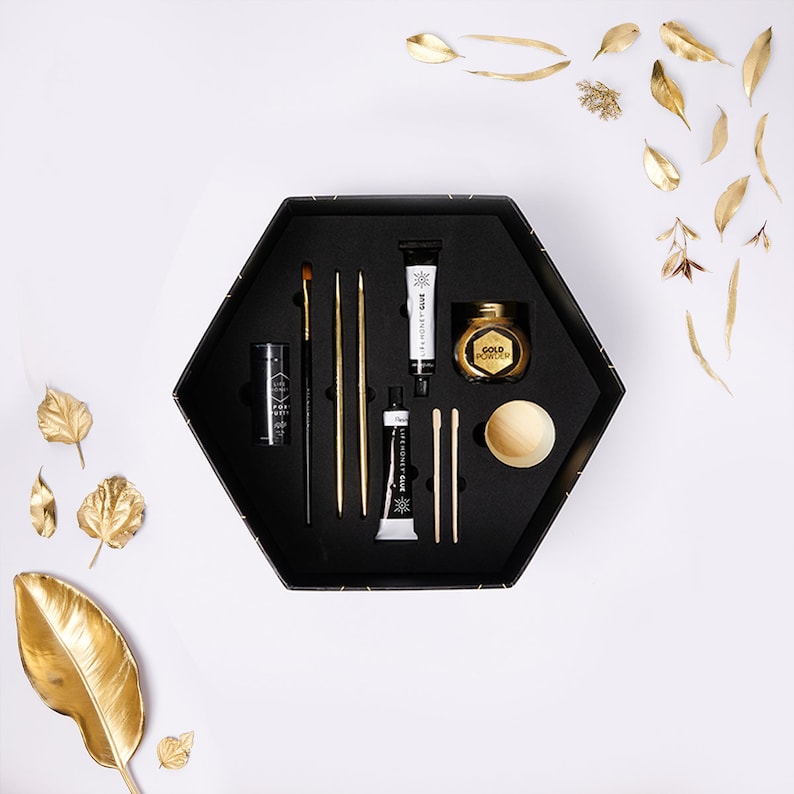 May include: A black hexagonal box with a variety of gold and black craft supplies, including paint, brushes, and a small jar of gold powder. The box is open and the supplies are arranged neatly inside.
