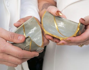 Puede incluir: Dos mitades de un cuenco de cerámica gris, unidas con detalles dorados, sostenidas por dos personas. Los bordes del cuenco están adornados en dorado. La imagen sugiere un tema de restauración o unidad, posiblemente para una boda o aniversario.