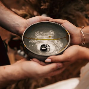 May include: A gray ceramic bowl with gold accents and two rings inside, held by two people. The bowl has a crack repaired with gold. A gold bracelet is visible on one wrist.