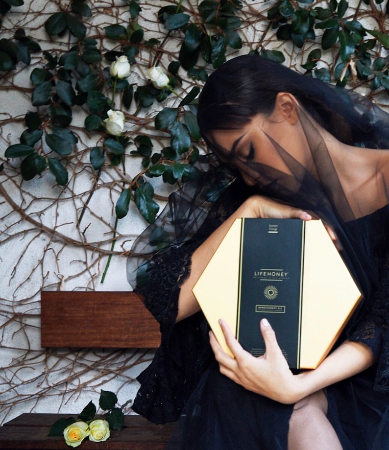 May include: A woman in a black dress and veil holds a hexagonal box with the text "LIFEHONEY" and a sun graphic. The box is gold with black accents.