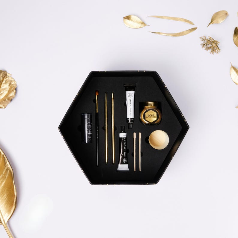 May include: A black hexagonal box filled with art supplies, including brushes, tubes of paint, and a small jar of gold powder. The items are arranged on a white surface with gold leaf accents, creating a visually appealing presentation for art enthusiasts.