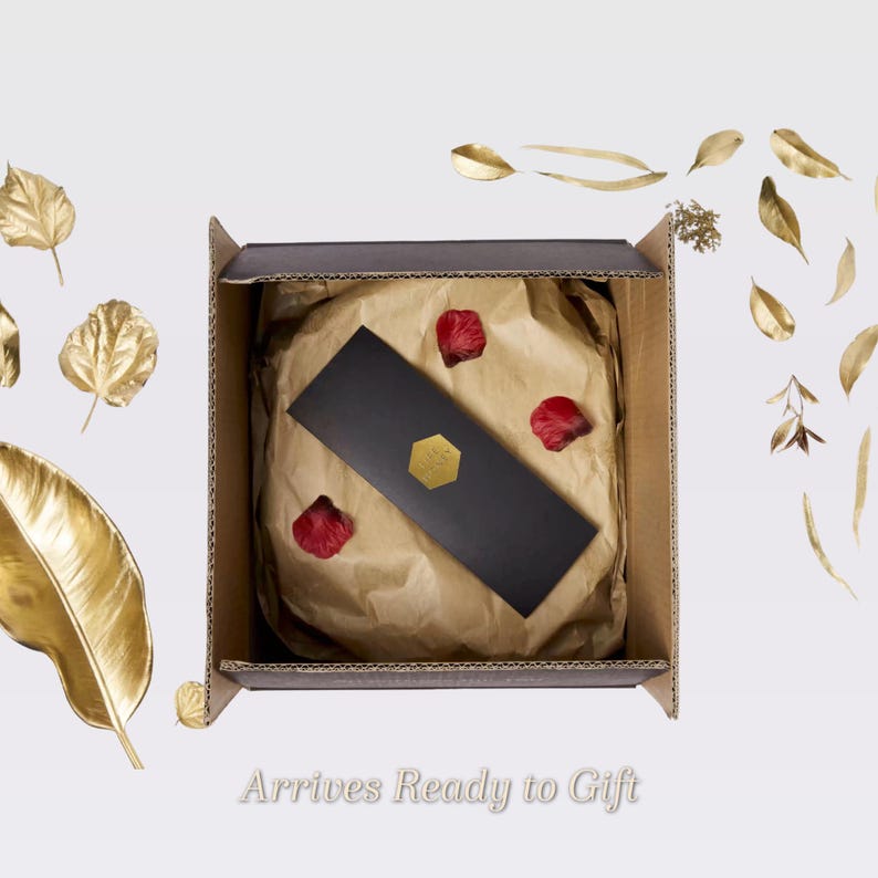 May include: An open cardboard box with a gift inside, lined with brown paper and adorned with red rose petals. A black rectangular card with a gold hexagon seal rests on top. Gold-colored leaves and the text "Arrives Ready to Gift" are also visible.