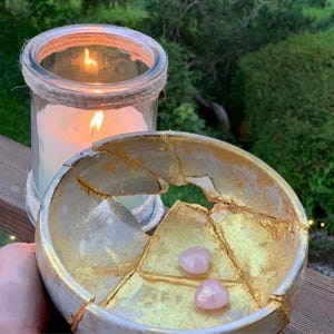 May include: A lit candle in a glass jar with a rope detail sits next to a broken bowl with gold accents and two small pink stones. The bowl has been repaired with gold leaf, showcasing the Japanese art of Kintsugi.