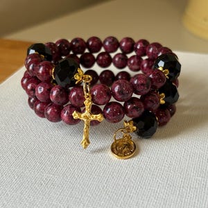 May include: A stack of three beaded bracelets. The bracelets feature dark red, round beads, interspersed with black, faceted beads. A gold-colored cross and a small charm hang from the bracelets.