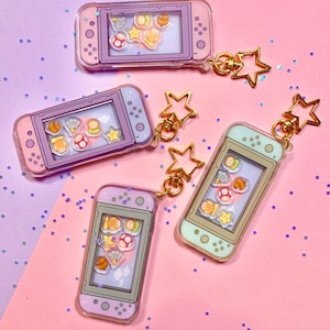 May include: Four colorful acrylic keychains shaped like game consoles, each with a gold star charm. The keychains feature various cartoon characters and shapes inside a clear, liquid-filled compartment. The consoles are in shades of purple, pink, and mint green.
