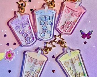 May include: Five colorful bubble tea charm keychains with gold star charms. Each keychain features a clear plastic cup with a straw and a variety of colorful boba pearls inside. The keychains are arranged on a pink and purple background.