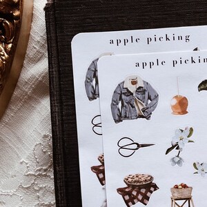 May include: Two sheets of white sticker paper with the text "apple picking" printed on them. The stickers feature illustrations of a denim jacket, a pair of scissors, a pie, a basket of apples, a ladder, and a blooming apple tree branch.