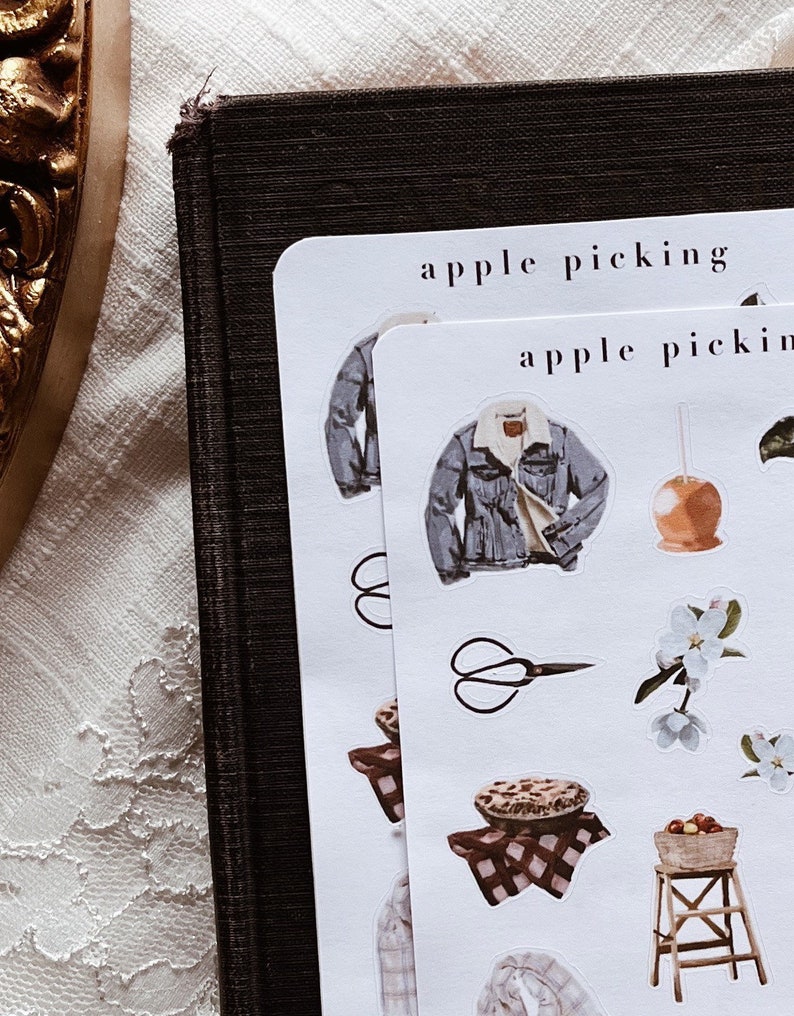 May include: Two sheets of white sticker paper with the text "apple picking" printed on them. The stickers feature illustrations of a denim jacket, a pair of scissors, a pie, a basket of apples, a ladder, and a blooming apple tree branch.