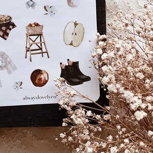 May include: A sheet of stickers with a variety of autumn-themed illustrations, including apples, boots, a cup of coffee, a basket of apples, a stool, and a flower. The stickers are on a white background and have a rustic, vintage aesthetic. The text "alwayslovelyco" is printed at the bottom of the sheet.