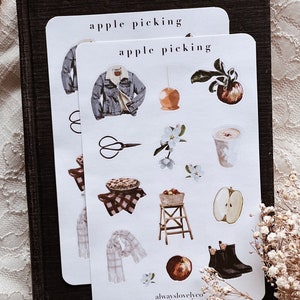 May include: A set of 12 apple picking themed stickers featuring illustrations of a denim jacket, a caramel apple, a pie, a pair of scissors, a basket of apples, a ladder, a scarf, a cup of coffee, an apple, boots, and a branch of apple blossoms. The stickers are printed on white paper and have a matte finish. The text "apple picking" is printed at the top of the sheet. The text "alwayslovelyco" is printed at the bottom of the sheet.