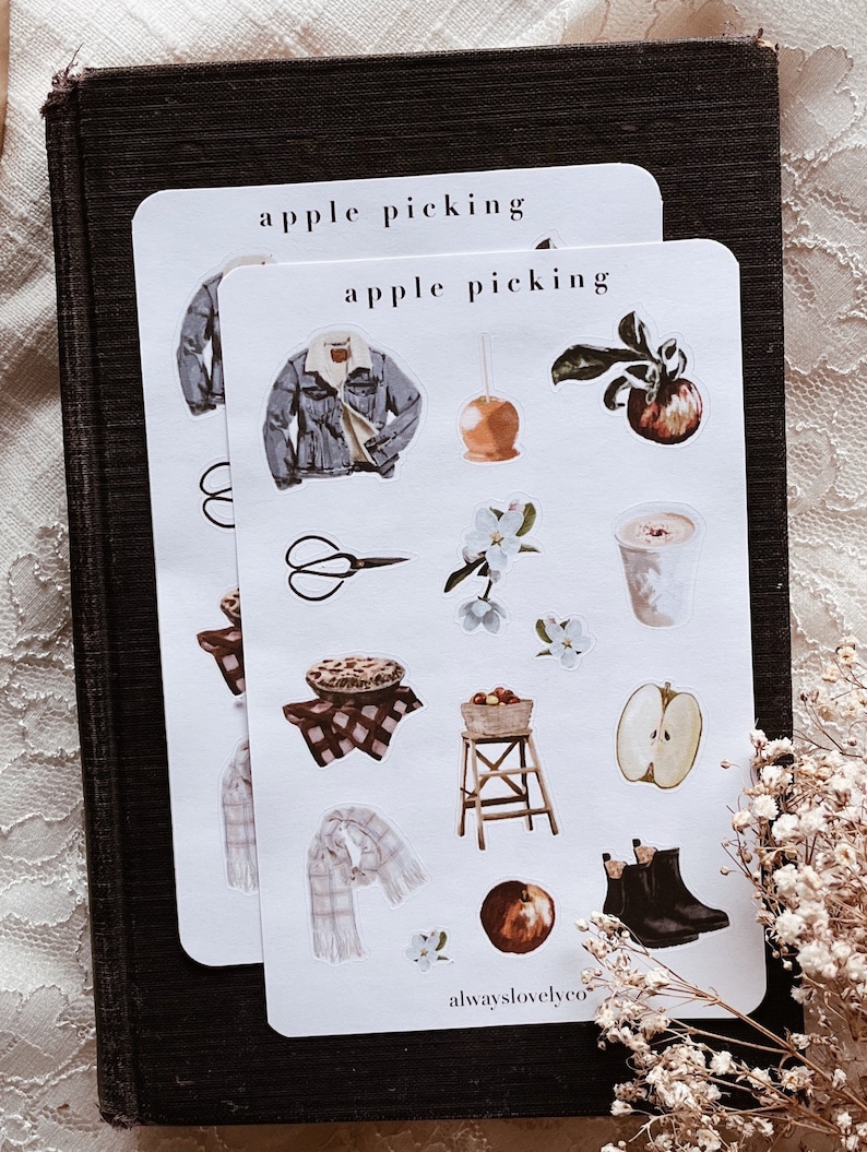 May include: A set of 12 apple picking themed stickers featuring illustrations of a denim jacket, a caramel apple, a pie, a pair of scissors, a basket of apples, a ladder, a scarf, a cup of coffee, an apple, boots, and a branch of apple blossoms. The stickers are printed on white paper and have a matte finish. The text "apple picking" is printed at the top of the sheet. The text "alwayslovelyco" is printed at the bottom of the sheet.