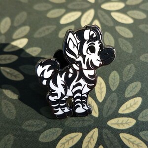 May include: A black and white enamel pin of a cartoon zebra with a black and white striped body and a white face. The pin is on a green background with a leaf pattern.