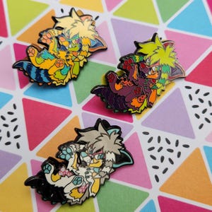 May include: Three enamel pins featuring cartoon animal characters. The pins are colorful and feature a variety of colors, including yellow, green, purple, orange, black, white, and blue. The pins are designed in a cute and playful style.