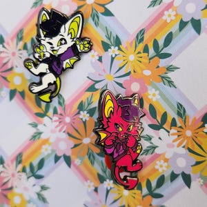Op de afbeelding: Twee emaillepins met cartoon-dierenfiguren met vleugels. De eerste pin is een wit personage met gele accenten en paarse kleding. De tweede pin is een rood personage met gele accenten en paarse kleding.