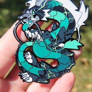 May include: A green and white enamel pin of a dragon with a fierce expression. The dragon is curled up with its tail in its mouth. The pin has a black outline and a shiny finish.