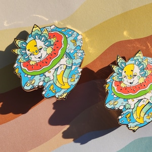 Könnte beinhalten: Zwei Emaille-Pins mit einem Cartoon-Axolotl, das ein Wassermelonen-Schwimmring trägt, in einer blauen und grünen Ozeanszene mit gelben Fischen.