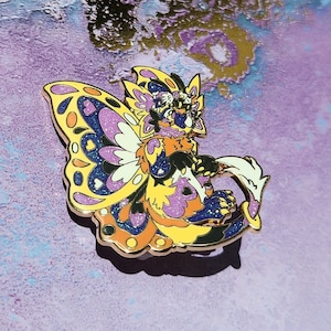 Puede incluir: Un colorido pin de esmalte con una criatura de fantasía con alas de mariposa. El pin tiene un contorno dorado y está decorado con purpurina amarilla, naranja, morada y azul. La criatura tiene un cuerpo peludo y un diseño caprichoso.