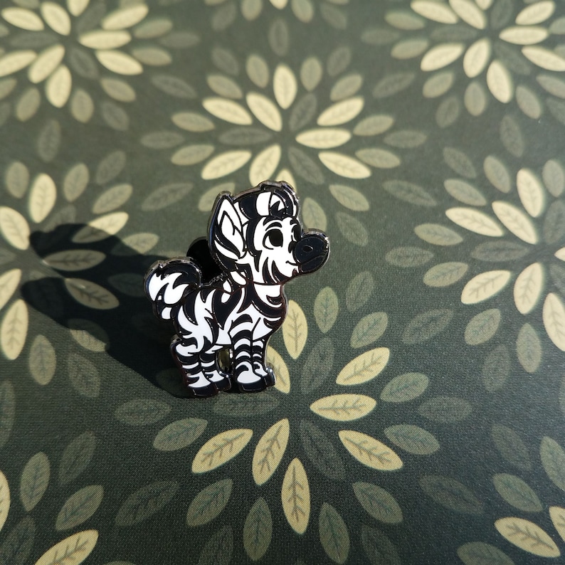 May include: A black and white enamel pin of a cartoon zebra with a black and white striped body and a white face. The pin is on a green background with a leaf pattern.