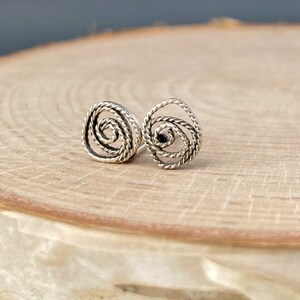 May include: A pair of silver stud earrings. Each earring has a spiral design made from twisted metal, giving a textured, openwork look. The earrings are shown on a light-coloured wooden surface.