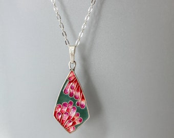 Può includere: Una collana d'argento con un ciondolo a forma di goccia. Il ciondolo presenta uno sfondo verde acqua con un disegno di forme floreali rosa e rosse. La catena della collana è delicata e presenta un motivo strutturato.