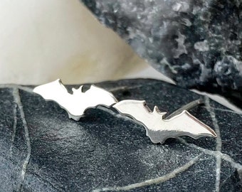 May include: A pair of silver bat-shaped earrings. The earrings are displayed on a dark gray stone, with a white and a gray stone in the background. The earrings have a simple, elegant design, perfect for Halloween or everyday wear.