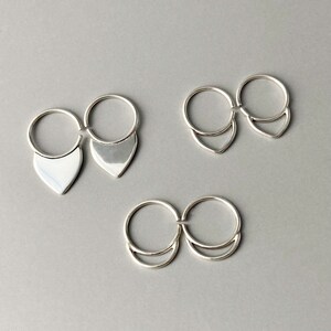 May include: Three pairs of silver rings. Each ring has a circular band connected to a decorative element. One pair features a teardrop shape, another a V-shape, and the last a crescent shape. The rings are displayed on a gray surface.