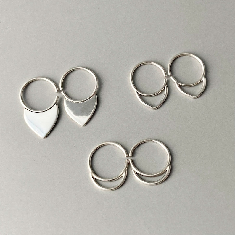 May include: Three pairs of silver rings. Each ring has a circular band connected to a decorative element. One pair features a teardrop shape, another a V-shape, and the last a crescent shape. The rings are displayed on a gray surface.