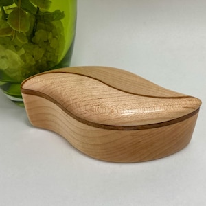 May include: A light-coloured wooden box with a smooth, curved, leaf-like shape and a lid that features a distinct, dark-toned inlay line running across the top surface, showcasing a natural wood grain pattern throughout the entire object.