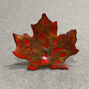 May include: A handcrafted, enameled maple leaf pin. The pin features a vibrant red base with intricate patterns of green and gold, creating a textured, autumnal appearance. The edges are outlined in a darker red, adding depth to the design.