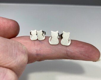 May include: Two pairs of silver cat-shaped stud earrings. One pair is smaller than the other. The earrings are displayed on a finger. The cats are facing away, with their tails curled. The earrings have a matte finish.