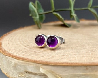 May include: A pair of round stud earrings with vibrant purple gemstones set in a silver bezel. The earrings are displayed on a light-colored wooden surface, with a sprig of green foliage in the background.