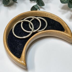 Puede incluir: Una bandeja para joyas en forma de media luna con exterior dorado e interior de terciopelo negro. Tres anillos de plata descansan dentro de la bandeja. La bandeja es de madera y tiene un acabado liso y pulido. El fondo es blanco con follaje verde.